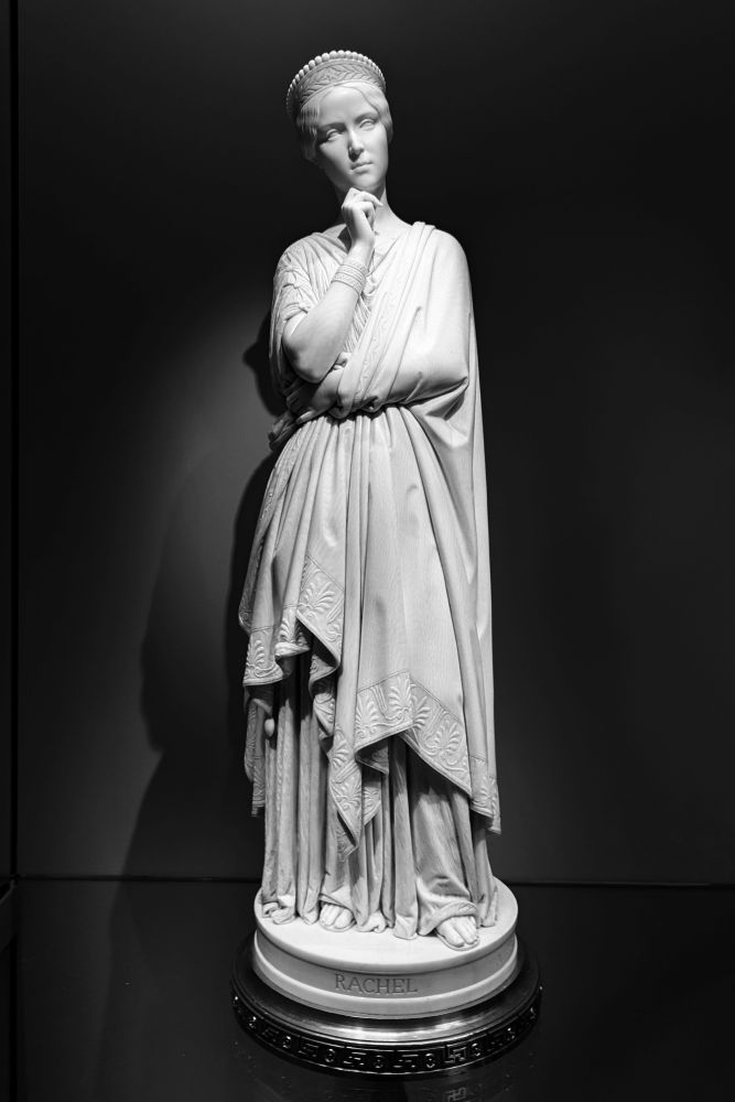 Rachel porte une robe qui la couvre entièrement.
Ses orteils dépassent des plis de la robe.
Son bras gauche est replié et serré son ventre.
Son bras droit prend appui sur sa main gauche pour soutenir son menton.
Rachel est pensive, le visage légèrement inclinée sur la droite.
Le revers du tissu laisse deviner un travail ouvragé. 
Rachel porte des bracelets au poignet droit et une tiare sur le front.