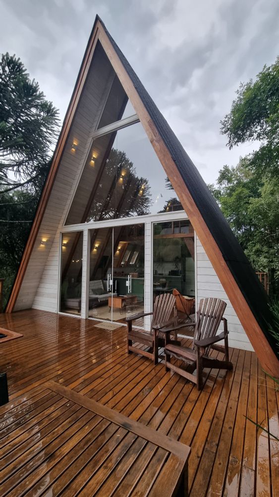 Foto da área externa mostrando uma cabana branquinha de madeira em formato de A rodeada por árvores. A parte da frente é toda de vidro, tem um deck de madeira onde é possível ver duas cadeiras de descanso. O céu está nublado