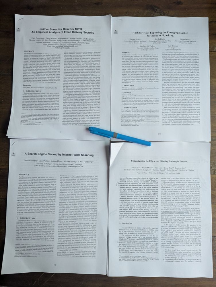 four printed research papers are laid on a table with a blue highlighter pen. The titles are "Neither Snow Nor Rain Nor MITM...An Empirical Analysis of Email Delivery Security," "Hack for Hire: Exploring the Emerging Market for Account Hijacking," "A Search Engine Backed by Internet-Wide Scanning," and "Understanding the Efficacy of Phishing Training in Practice"