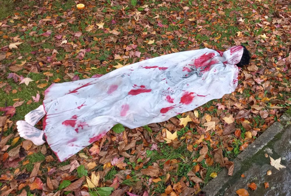 Un cadáver en un jardín lleno de hojas. Está cubierto por una sábana ensangrentada y le sobresalen los pies. 