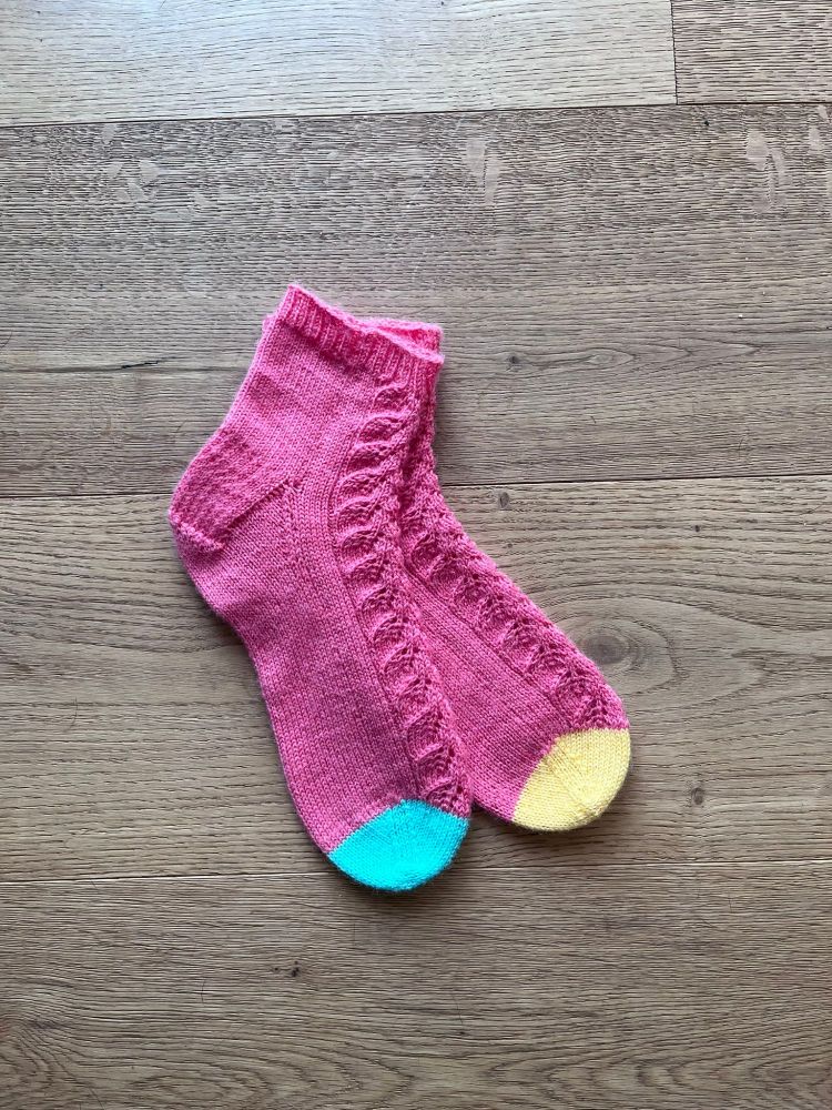 a pair of pink socks displayed flat with a leaf lace pattern going down the front of each sock, with one contrast toe in green and another in yellow