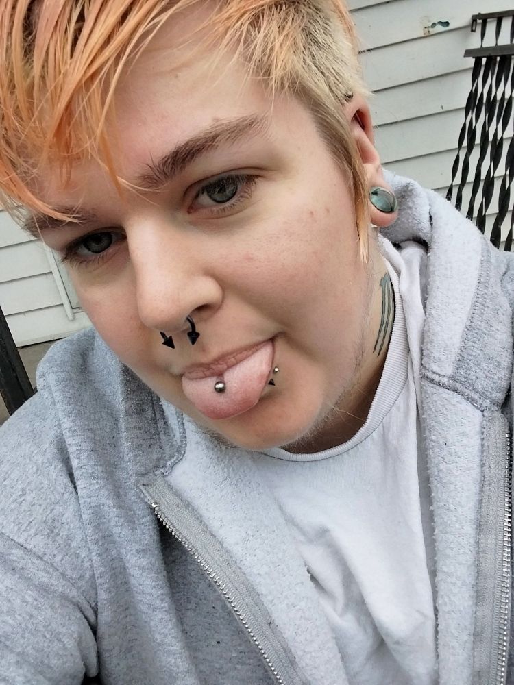 Selfie of a Nonbinary person with scraggly beard and orange-blonde hair with shaved sides, glass gauges, several face piercings, sticking their tungue out facing the camera 