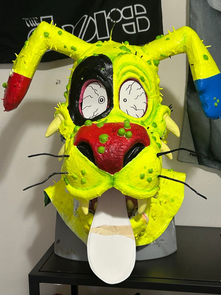 The photo depicts the progress of a of a dog mask in a cartoony grotesque style. The mask is neon yellow/green with blue and red ears. The dog has a red nose and black whiskers. He is covered in green acne. 