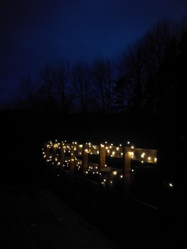 Bilde av lysende led-lyslenke, hengt opp langs rekkverk, i mørket. Skimter trær og blåsvart himmel i bakgrunn