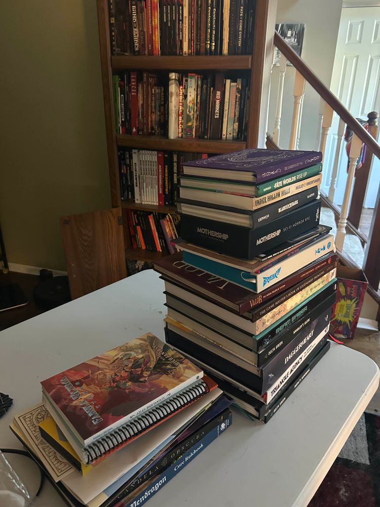 A photo of two piles of tabletop roleplaying games. Behind them is a bookshelf, which is ALSO full. (Yes, even the two shelves you can’t see below camera’s edge.)