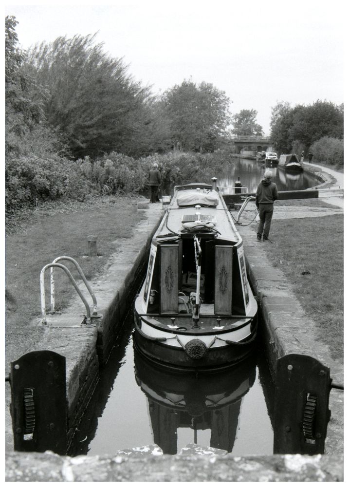 A narrow boat going up up a lock, looking from a bridge behind the lock, there's a small terrier dog by the tiller. Photo is black and white. 