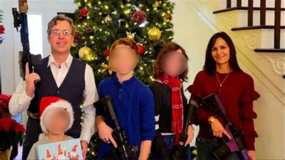 MAGA terrorist Andy Ogles poses with his wife and three children in front of a Christmas tree. Ogles, his wife, and the two oldest children hold assault rifles, while the youngest child holds a wrapped present. The faces of the children have been blurred, Ogles and his wife have looks of pride and contentment on their faces. 