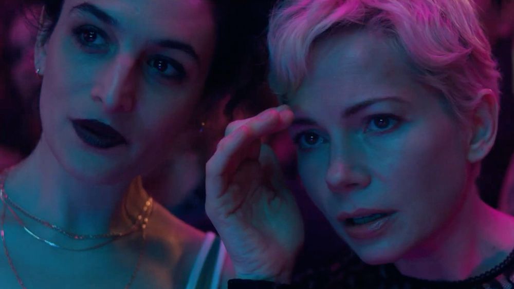 Michelle Williams and Jenny Slate stand side by side in pink lighting, Jenny Slate with dark lipstick and jewelry like she's ready for a club night and Michelle Williams dressed casually. They have their heads together talking about something off screen, Williams shielding her face like she's hiding. They look intent but lightly amused.