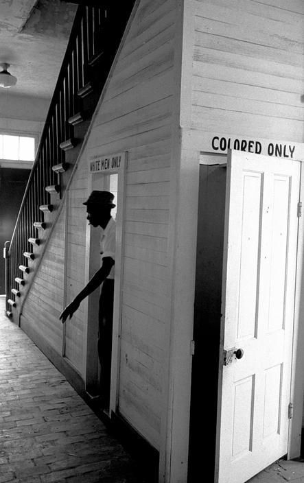 a man in a hat emerges from a doorway situated beneath a large set of stairs