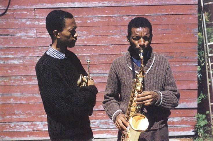 phito of two men standing outside near a red building with clapboard wood siding. the man on the left is don cherry, wearing a black sweater with arms crossed holding a trumpet. he is faces the second man who wears a brown sweater, is facing straight ahead, and blowing a saxophone.
