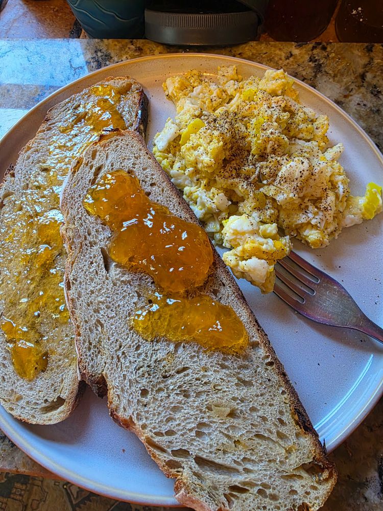 photo of 2 slices of sourdough bread covered in peach preserves next to scrambled eggs on a white plate next.