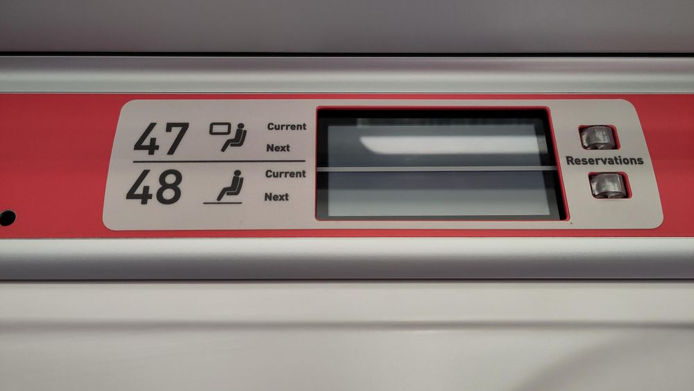 Signs in the interior of a train: on physical signage, the numbers 47 and 48 next to icons depicting seated humans. Next to that are two liquid crystal displays, which are blank. Next to that, two small lights with the word "Reservations", neither light is illuminated 