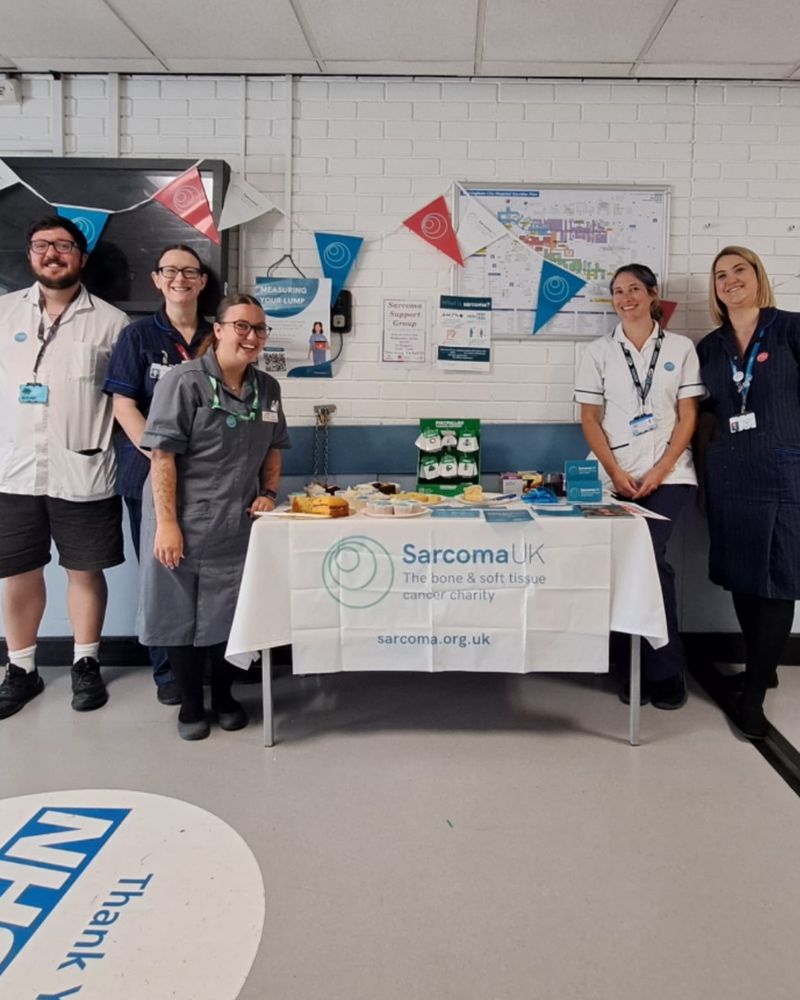 Photo of the hospital team standing around the awareness stand which has cakes on and a Sarcoma UK table cloth. Sarcoma UK bunting hangs in the background.