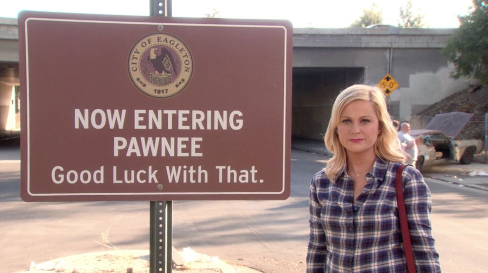 Leslie standing in front of a sign that says Now Entering Pawnee Good Luck With That