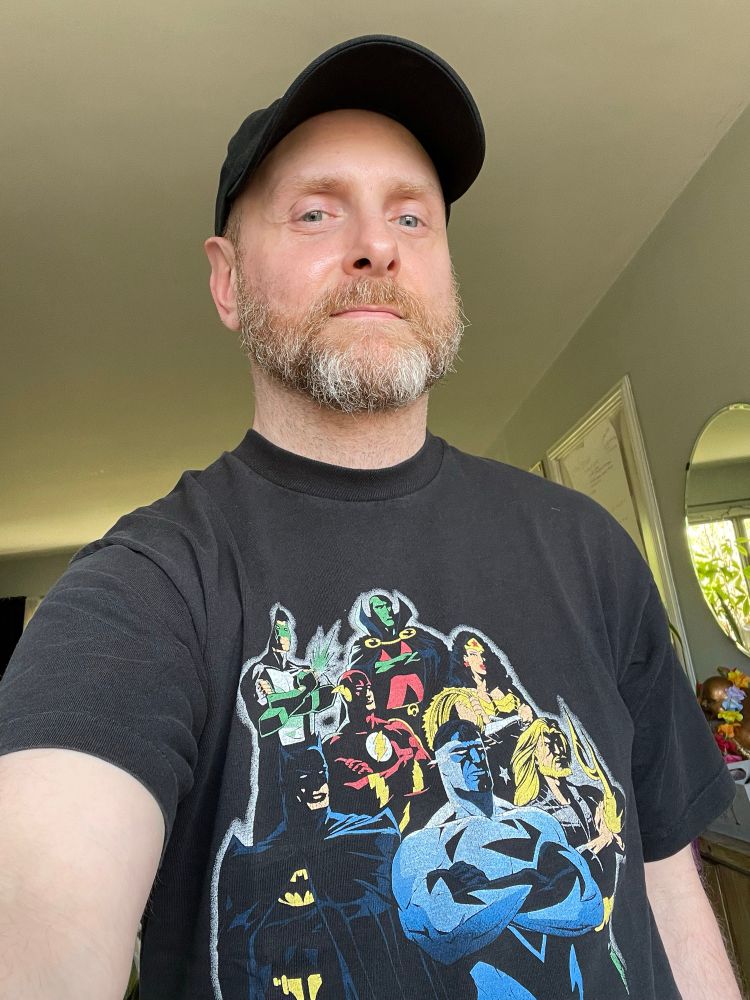 Me wearing a black baseball cap and a JLA tee shirt featuring electric superman, hook hand Aquaman, Wonder Woman, Batman, green lantern Kyle rayner, the flash and Martian Manhunter 