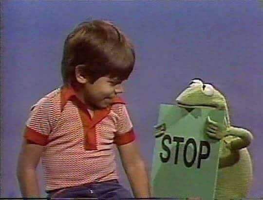 Kermit the Frog holding “STOP” sign in front of white boy wearing red shirt. 