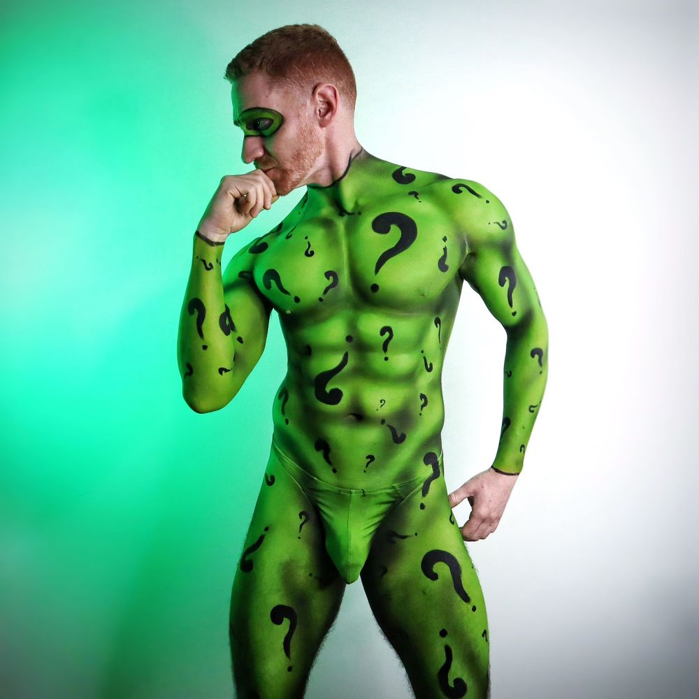 Studio shot of Leander, an athletic redhead, who has been body painted to look like the riddler. He is all green from the neck down, with questions marks all over him and heavily accented lines emphasising his muscular body.