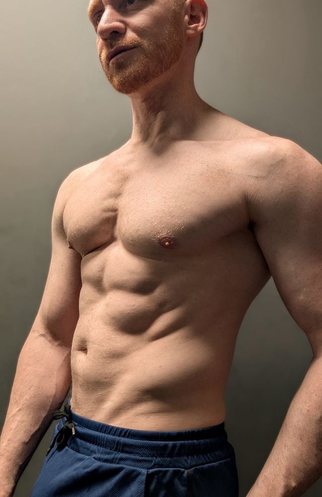 Cropped close up selfie of redhead male Leander’s torso with strong down lighting emphasising his muscles.