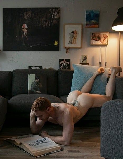 Leander, an athletic male redhead, is lying naked, face down, half on, half off, a sofa. His arms are resting on the ground but his pelvis is resting on the sofa and his legs extend up one arm of the sofa. He is reading a book that's lying open on the floor and the position makes an arch in his back that accentuates his muscular ass.