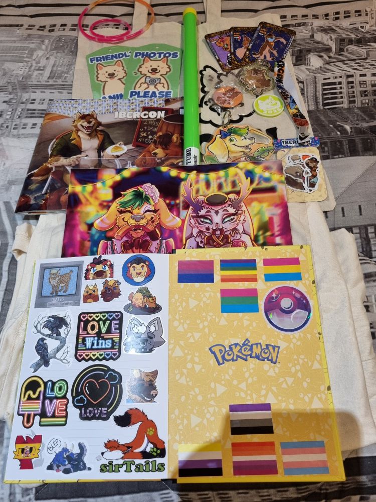 A photo that shows all the cool merchandise that I got at Ibercon 2025 🤗.
There are lots of stickers, a room sign done by my best friend Odei that shows both of our sonas eating churros, two tags that say photos please and friendly animal, two tshirts, a badge that shows my sona Hawaii wearing a tropical tshirt and drinking a cocktail, made by Lionrae, three themed cards of the convention, a bag that shows the conventiom mascots Adri and Bobi, a dog and an iberiam wolf, a pin that features a chibi Bobi, the conbook, a green neon stick that makes a nyaan noise, two neon brazelets and one soft brazelet and a neon green coin that features Adri.