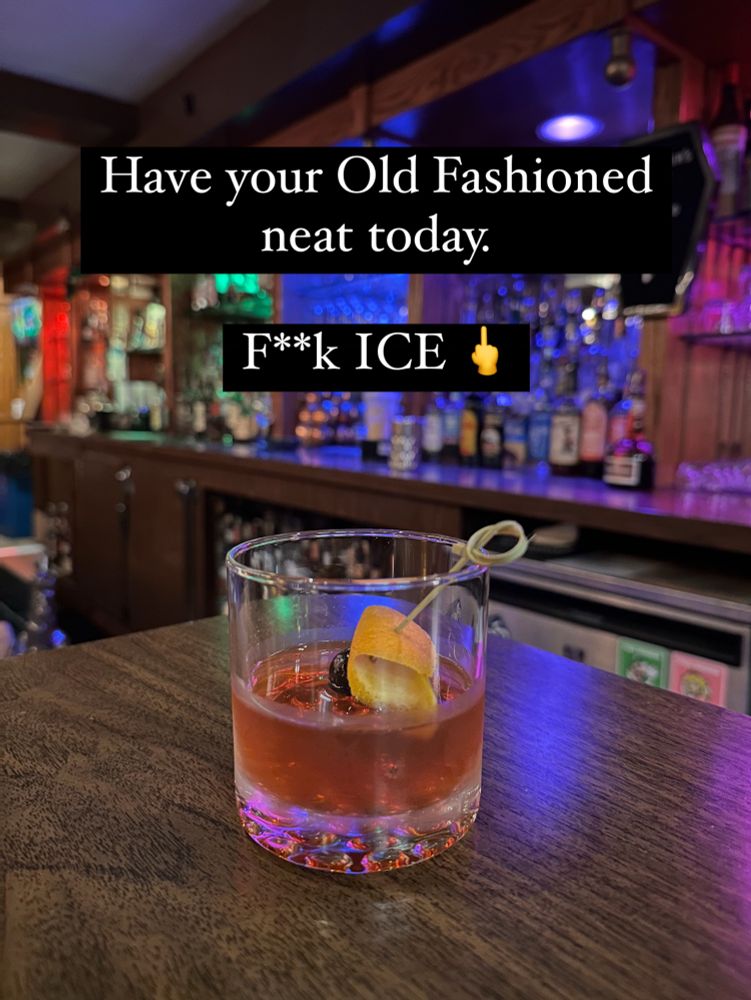 An Old Fashioned cocktail with NO ICE sits on a bar with Pride lighting behind