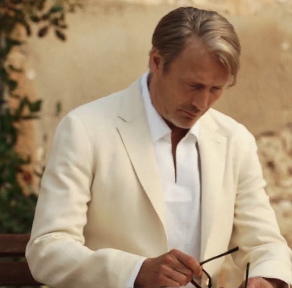 Actor Mads Mikkelsen is wearing a cream linen jacket and white shirt. He holds a pair of glasses in his right hand. There is a cream wall behind him, and a tall plant in a terracotta pot. He seems to be outside. He is looking serious.