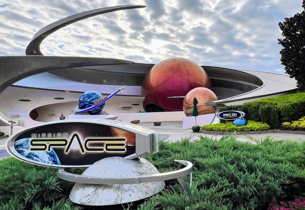 The ride sign for Mission: Space sits under an overcast sky. The sign uses a future spacey looking font and has images of both Earth and Mars behind the text, with the sign itself protruding from half of a white sphere, possibly to represent the Moon. A large circular metal arm comes out of the sign to circle the Moon piece.

Similar arms in much larger scale make up the ride’s facade, visible behind the sign. They circle around three full globes: one blue and white to represent Earth, one dark red to represent Mars, and a third smaller orange one, which is like second Mars? A little miniature spaceship is leaving the Earth globe towards the Mars one on a blue arm.

Trimmed bushes and plants surround both the sign and the facade.