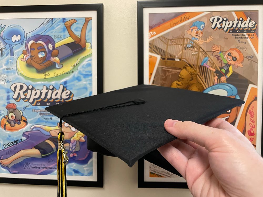 Holding a 2025 undergrad graduation cap in front of posters of the LAN events Riptide 2022 and Riptide 2023