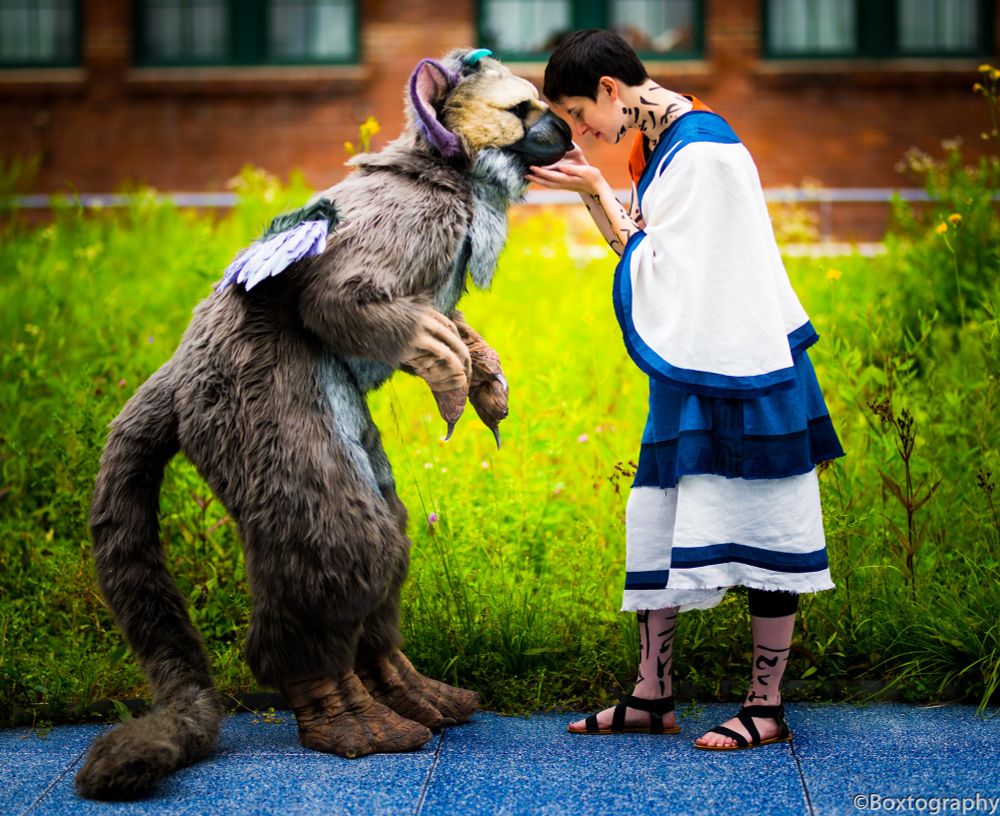 A cosplay photo of a person holding a beast's chin with their forehead to its beak. The beast is leaning forward in cautious curiosity.
