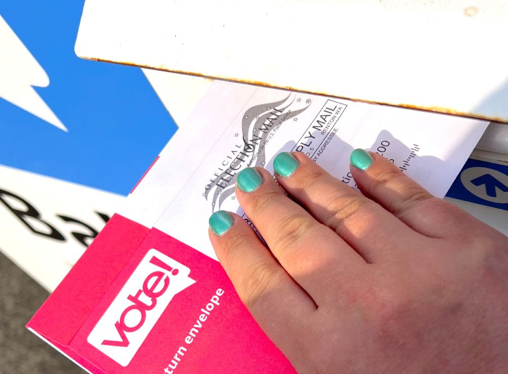 A man's hand with short fingernails and bright teal green nail polish deposits a vote-by-mail ballot into a public ballot collection box.