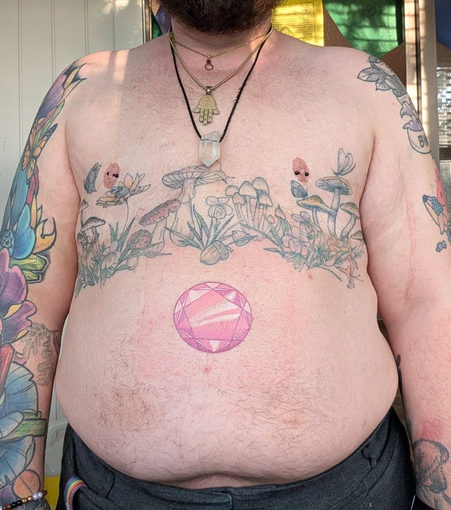 A shirtless nonbinary person (fae/they) with tattoos. A mushroom and plant chestpiece with small critters (a buttefly and bee are noticeable), floral and crystal sleeve on faer right arm, and some other tattoos not very visible on faer left arm. Below the chest piece on faer belly is a Steven Universe-esque rose quartz gem in a more realistic style. Fae is also wearing three stacked necklaces; the nonbinary symbol, a hamsa, and a quartz crystal.