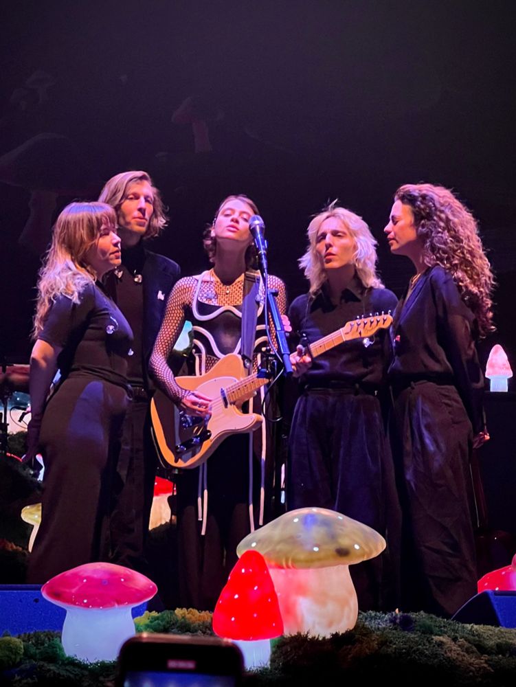 Pomme chantant en chœur avec ses musiciens de scène en fin de concert, réunis en demi-cercle autour d'un seul micro. 

Pomme tient une guitare électrique et de petits champignons lumineux dominent le premier plan à leurs pieds.