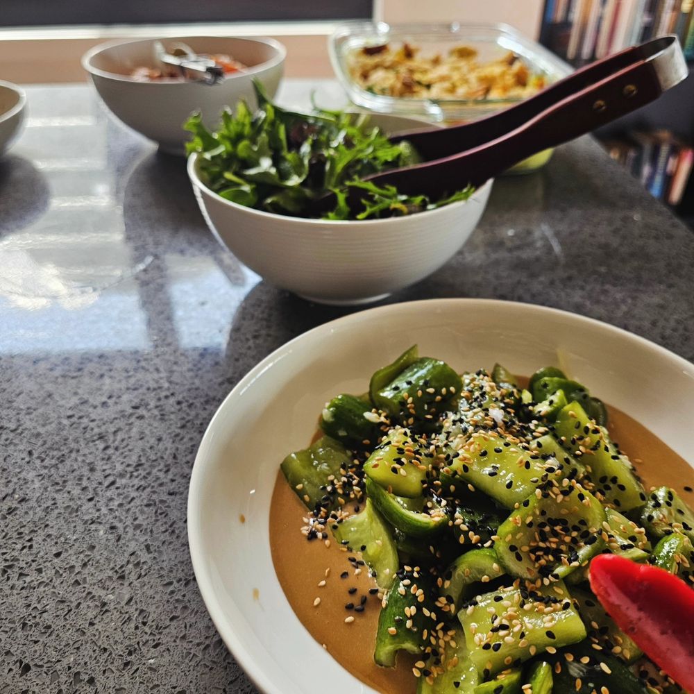 Lunch all plated up and ready to serve buffet style. In the foreground is tangy smashed cucumbers with Tahini & soy dressing; in the background is a bowl of green leaves, another of spiced honey carrots, and a cauliflower & sweet potato bake with curried cashew cream