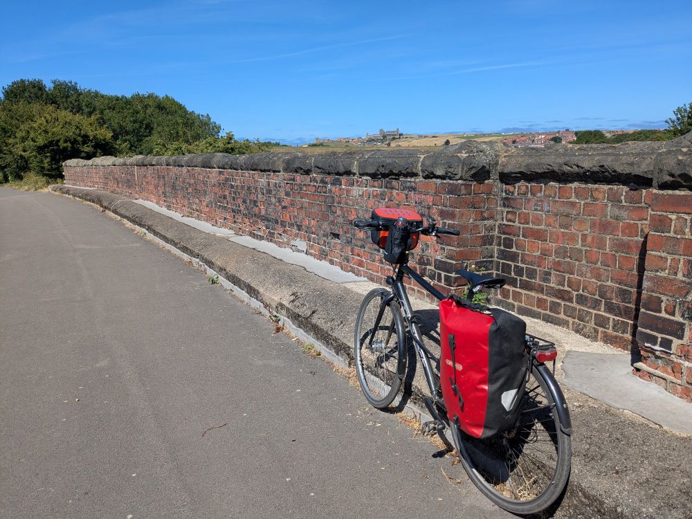 Touring bike leaning against the pavement on a bright sunny day, with wall of the Larpool Viaduct behind and Whitby Abbey in the background on the horizon
