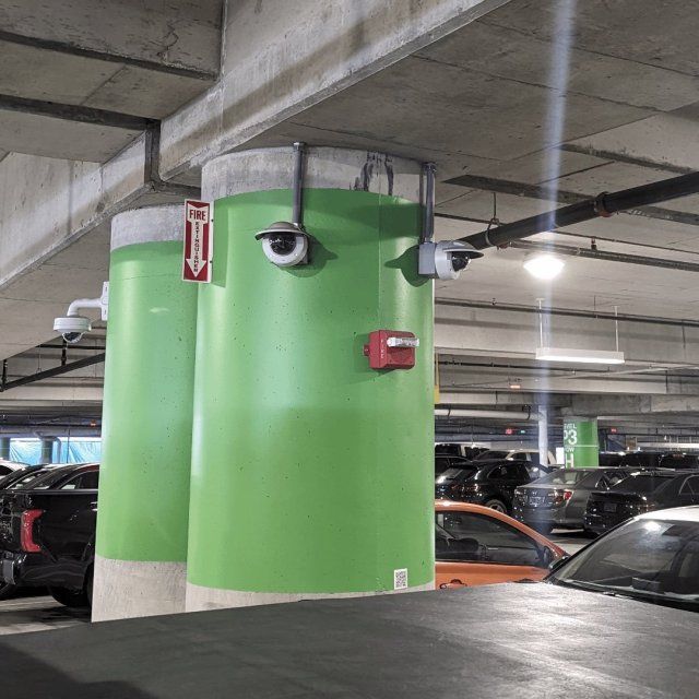 The image shows a parking garage with a green pillar. The pillar has been made to look like a face, using light fixtures or security cameras for the eyes and a red fire alarm box as the mouth. The expression created makes the pillar appear to have a surprised or concerned look.