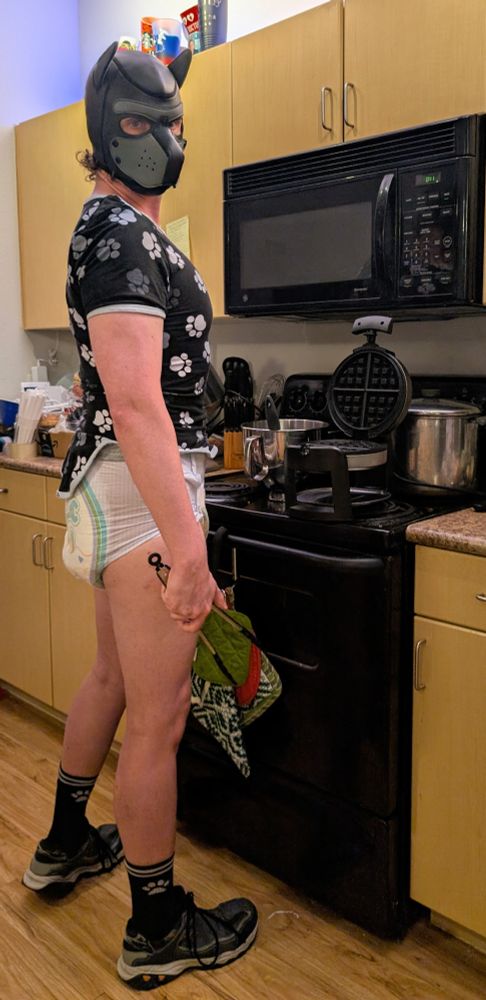 Human pup wearing a pup hood, matching paw print onesie, paw-print socks, and an alphagatorz diaper.  He's holding tongs and standing in front of a Belgian waffle press in a kitchen.