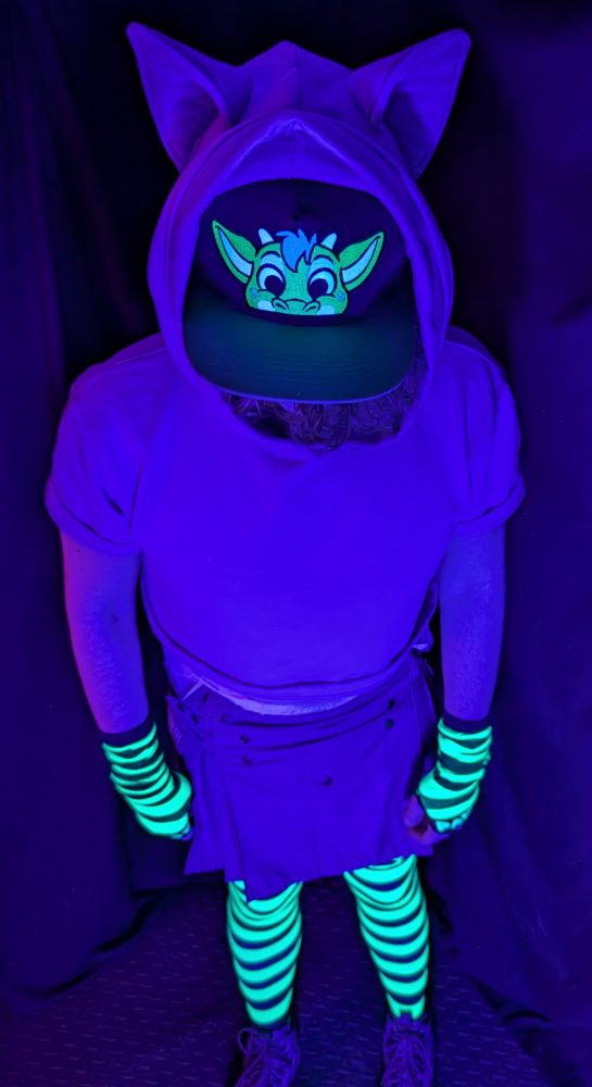Babyfur boyo in blacklight, while wearing a short, purple kilt, a purple crop top, a team Dragon (Derg) ABUniverse (ABU) Peek hat, green-and-black striped socks with matching arm warmers (from angypaws), and purple boots.  A DinoRawrz diaper rides up from the kilt into the tummy area between kilt and crop top. The hat's full color range is glowing well, along with the socks and arm warmers.