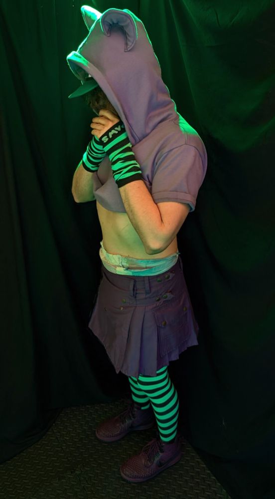 Babyfur boyo being bashful while wearing a short, purple kilt, a purple crop top, a team dragon ABU peek hat, green-and-black striped socks with matching arm warmers, and purple boots.  A DinoRawrz diaper rides up obviously from the kilt into the tummy area between kilt and crop top.  He's backlit by a green-colored PAR light.