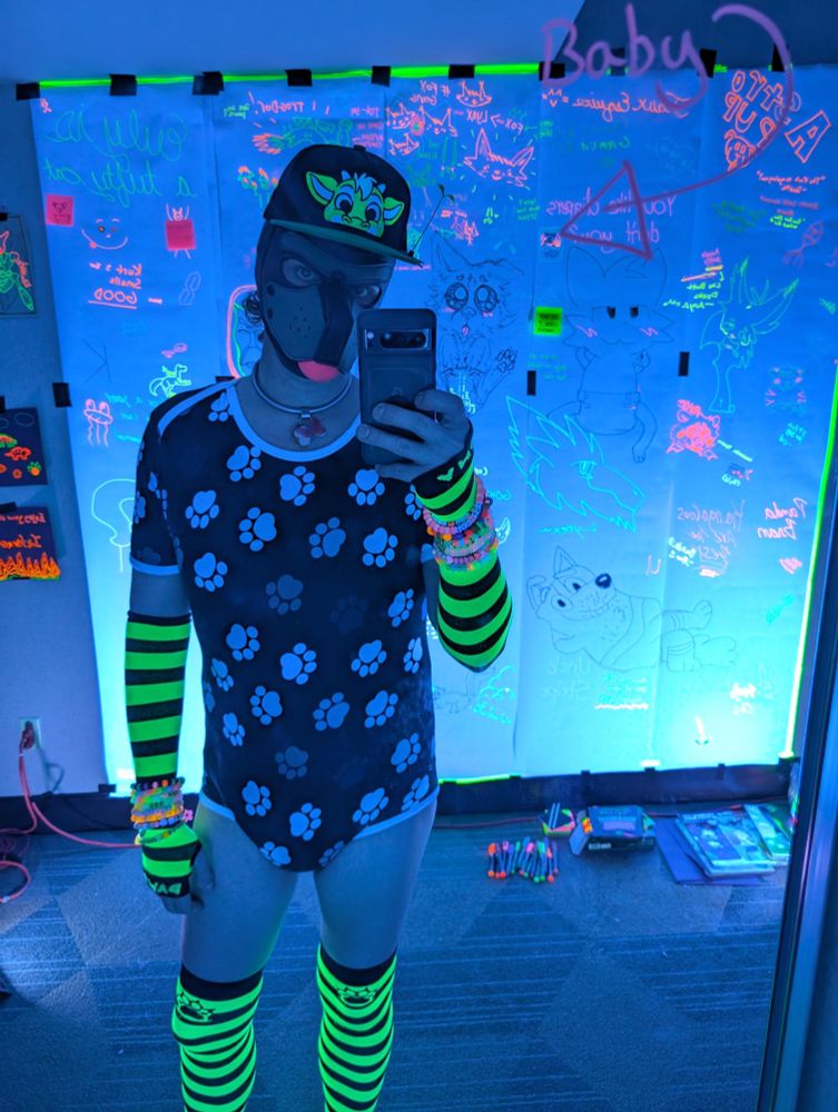 Babyfur taking a selfie in front of a mirror while wearing a pup hood with a neon-colored dragon peekabu hat on top.  Green and black striped socks and arm warmers. Wearing a puppy paw print onesie.

A large wall is behind him covered by paper and hand-written drawings and text, almost all of which are neon and illuminated by blacklights.