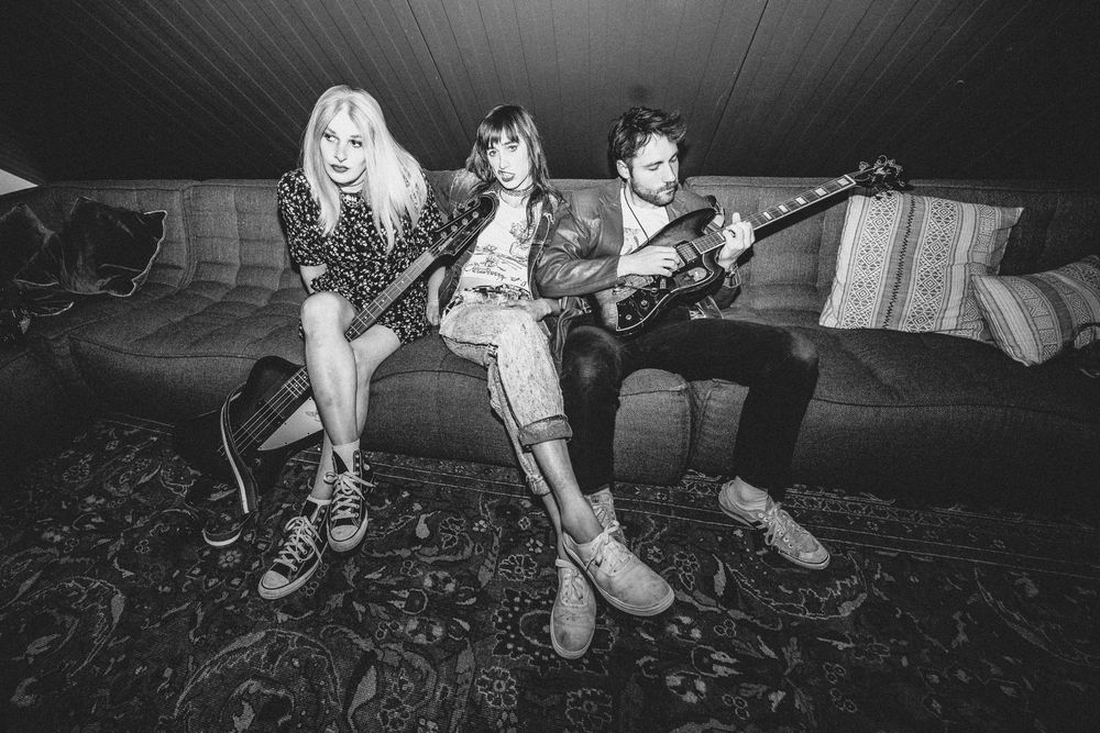 New black and white press photo of the band Ekko Astral by Murphy Gerrasch. Band members Jael Holzman, Miri Tyler and Liam Hughes all sit together on a couch backstage at one of the shows, guitars in hand.