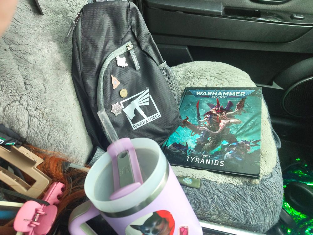 A photo of a car passenger seat covered in grey wool car covers.
Stop it sits a black "Warhammer" branded bag with some indistinct pins and a Warhammer Tyranids codex book.