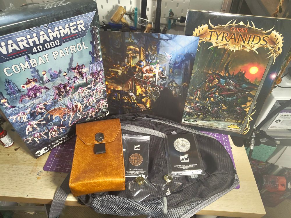 A photo of a series of Warhammer merchandise pieces on a desk.

Left to right:
A Warhammer 40,000 tyranid combat patrol, an art print of a space marine sitting on a throne, and an artwork of an old Tyranids codex print.
Bottom row: a leather Warhammer dice pouch, s bronze coin, a silver coin, two skull pins, all sitting in a black Warhammer sling bag.