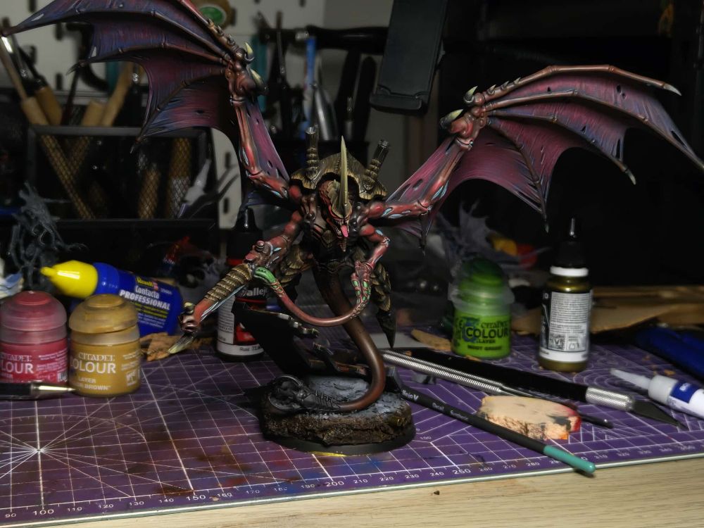 A Winged Hive Tyrant from Warhammer 40k's Tyranids. This front-on shot shows the deep red skin, the almost purple wings, and brown carapace. The base for the model has a ruined building wall corroded in browns, reds, and yellows.