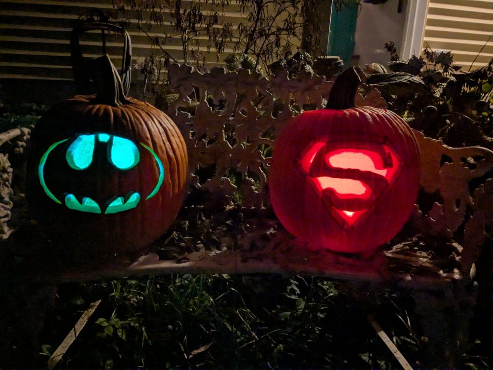 Two jack-o'-lanterns sitting on a painted iron bench in a dark yard. The porch light is on behind them. The pumpkin on the left has the Batman logo carved into it and is lit up blue. The pumpkin on the right has the Superman logo carved into it, and is lit up red.