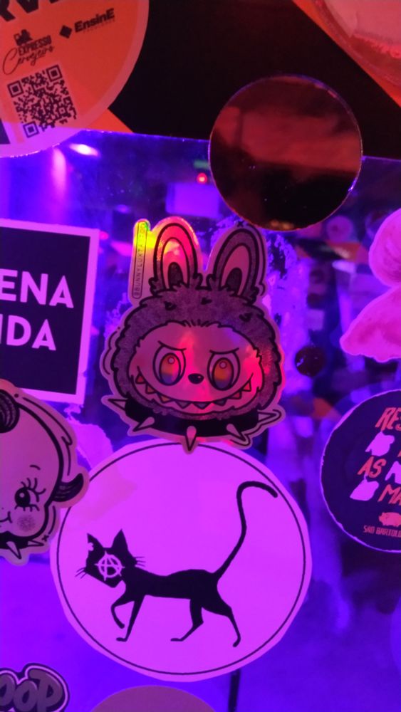 Foto de uma parede de vidro com vários adesivos. No centro da parede dá pra ver um adesivo de um labubu usando um colar de spike e os olhos na cor do arco íris