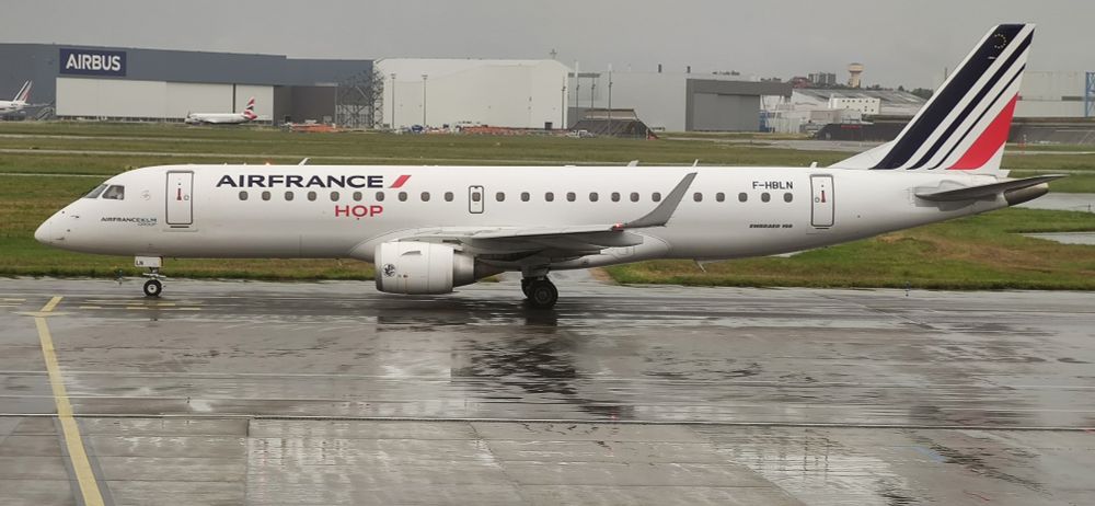 F-HBLN Emb190 of Air France Hop at Toulouse in May24. Aircraft is on a rain soaked ramp with grass behind and Airbus hangars in the distance