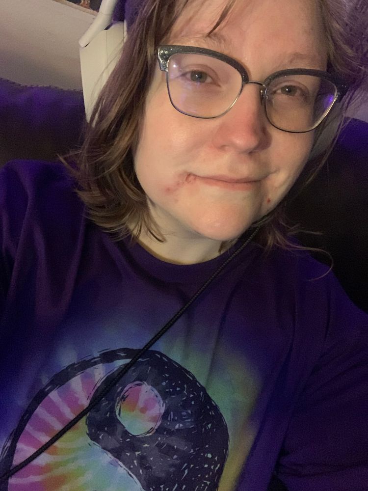 A selfie of TRRG. They’re wearing a purple shirt with the yin and Yang symbol on it, but the white is filled in with tie dye instead. They’re wearing their gray headphones and black framed glasses. They are smiling slightly 