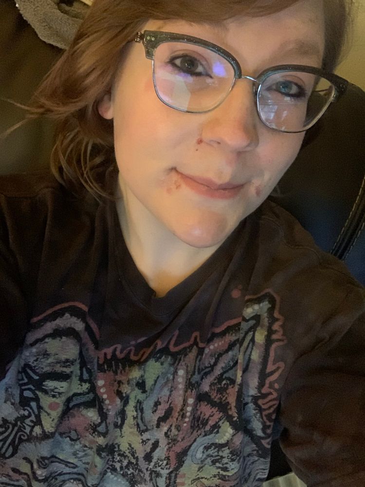 A selfie of TheRealRascallyGrandpa, wearing a t shirt that seems to have some abstract art on it. 

They are wearing eyeliner, mascara, and foundation, and smiling at the camera. Their hair is short and reddish and there is a glare in their glasses from screens. 