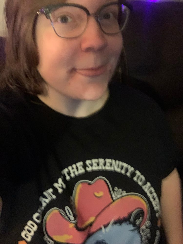 A slightly blurry selfie of TRRG, sticking their tongue out. They’re not wearing makeup. They’re wearing a black shirt that has a possum on it with a cowboy hat. The text is not fully visible. 