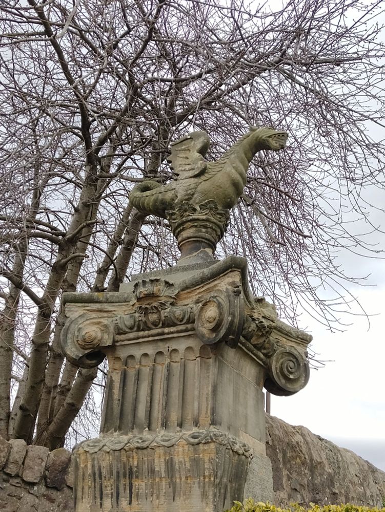 Concrete statue of a wyvern atop a squared, fluted Corinthian style column.